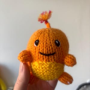 May include: A crocheted orange and yellow plush toy with a flame-shaped tail. The toy has black eyes and a smiling mouth.