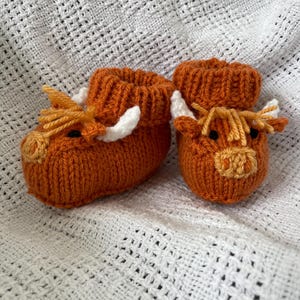 May include: A pair of knitted baby booties shaped like brown Highland cows with white horns and orange noses.