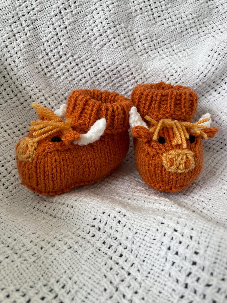 May include: A pair of knitted baby booties shaped like brown Highland cows with white horns and orange noses.