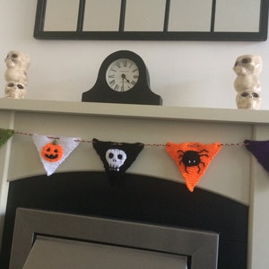 May include: A Halloween banner with five triangular flags. The flags are crocheted and feature different Halloween motifs, including a bat, a pumpkin, a skull, a spider, and a tombstone. The banner is strung across a white fireplace mantel.