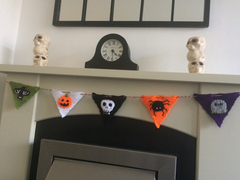 May include: A Halloween banner with five triangular flags. The flags are crocheted and feature different Halloween motifs, including a bat, a pumpkin, a skull, a spider, and a tombstone. The banner is strung across a white fireplace mantel.