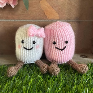 Può includere: Due giocattoli di peluche a forma di marshmallow con facce sorridenti. Il marshmallow a sinistra è bianco con un fiocco rosa e il marshmallow a destra è rosa. Entrambi sono seduti sull'erba verde.