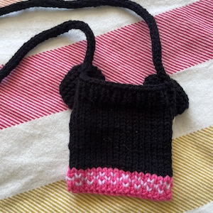 Pu&ograve; includere: Una borsa di maglia nera con bordatura rosa. La borsa ha una lunga tracolla ed &egrave; abbastanza piccola da stare in tasca.