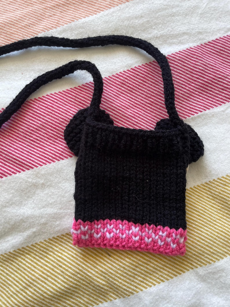 Pu&ograve; includere: Una borsa di maglia nera con bordatura rosa. La borsa ha una lunga tracolla ed &egrave; abbastanza piccola da stare in tasca.