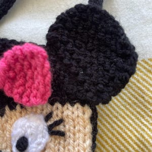 Pu&ograve; includere: Una borsa a mano di maglia a forma di personaggio dei cartoni animati con orecchie nere, un fiocco rosa e un occhio bianco. La borsa &egrave; beige con ciglia nere.