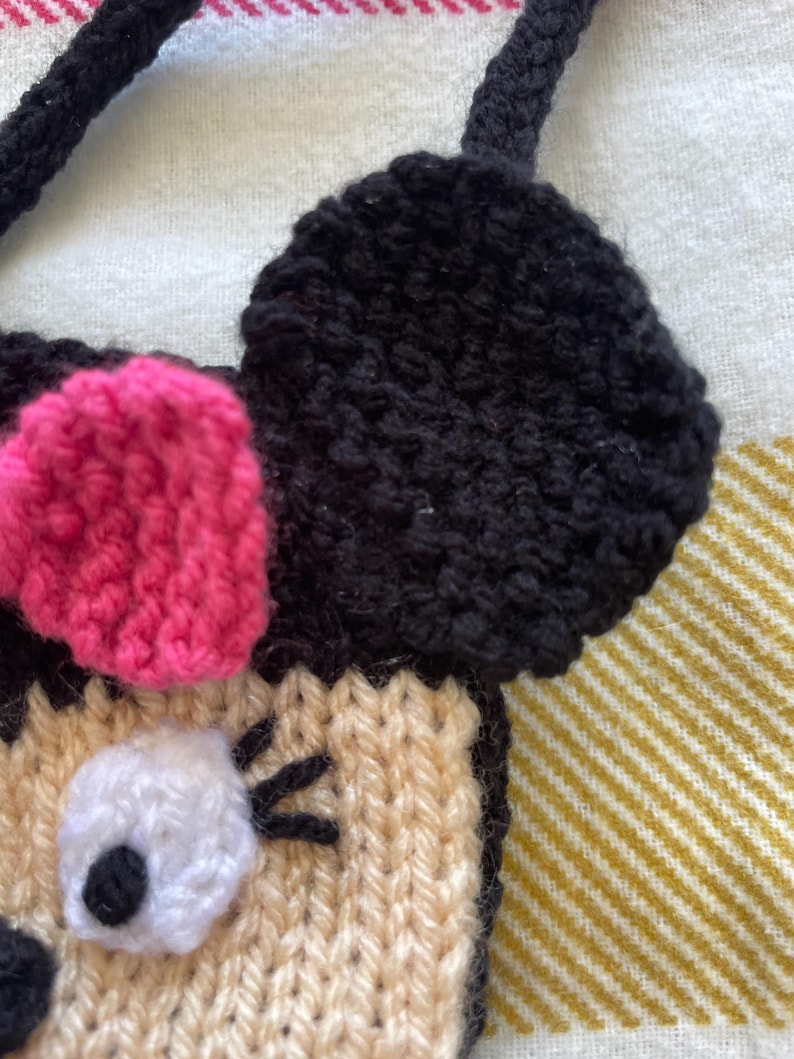Pu&ograve; includere: Una borsa a mano di maglia a forma di personaggio dei cartoni animati con orecchie nere, un fiocco rosa e un occhio bianco. La borsa &egrave; beige con ciglia nere.