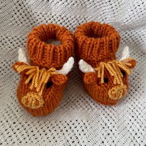 May include: A pair of knitted baby booties with a brown highland cow design. The booties have white horns, a brown face, and a yellow nose.