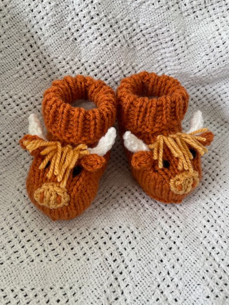 May include: A pair of knitted baby booties with a brown highland cow design. The booties have white horns, a brown face, and a yellow nose.