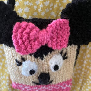 Pu&ograve; includere: Una borsa a mano di maglia a forma di personaggio dei cartoni animati con orecchie nere, un fiocco rosa e un viso bianco con occhi neri e un naso nero. La borsa ha un bordo a righe rosa e bianche.