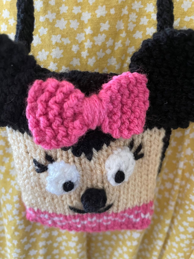 Pu&ograve; includere: Una borsa a mano di maglia a forma di personaggio dei cartoni animati con orecchie nere, un fiocco rosa e un viso bianco con occhi neri e un naso nero. La borsa ha un bordo a righe rosa e bianche.