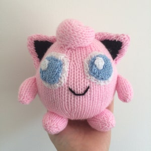 May include: A pink knitted plush toy resembling a Pokemon character with black ears, blue eyes, and a white smile.