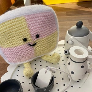 Può includere: Un giocattolo di maglia a forma di quadrato con quadrati rosa e gialli, con una faccia felice, seduto a un tavolo bianco con pois neri, accanto a una teiera bianca con pois neri e un creamer bianco con pois neri.