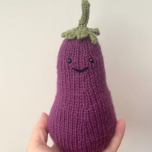 Puede incluir: Un juguete de peluche de berenjena tejido a mano. El peluche es de color morado oscuro con un tallo y hojas verdes. Tiene una cara sonriente con ojos negros y una boca curvada. El juguete se sostiene en una mano.