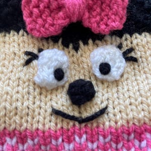Pu&ograve; includere: Un cappello di lana con un fiocco rosa, orecchie nere e un viso da cartone animato con occhi bianchi, un naso nero e una bocca sorridente.