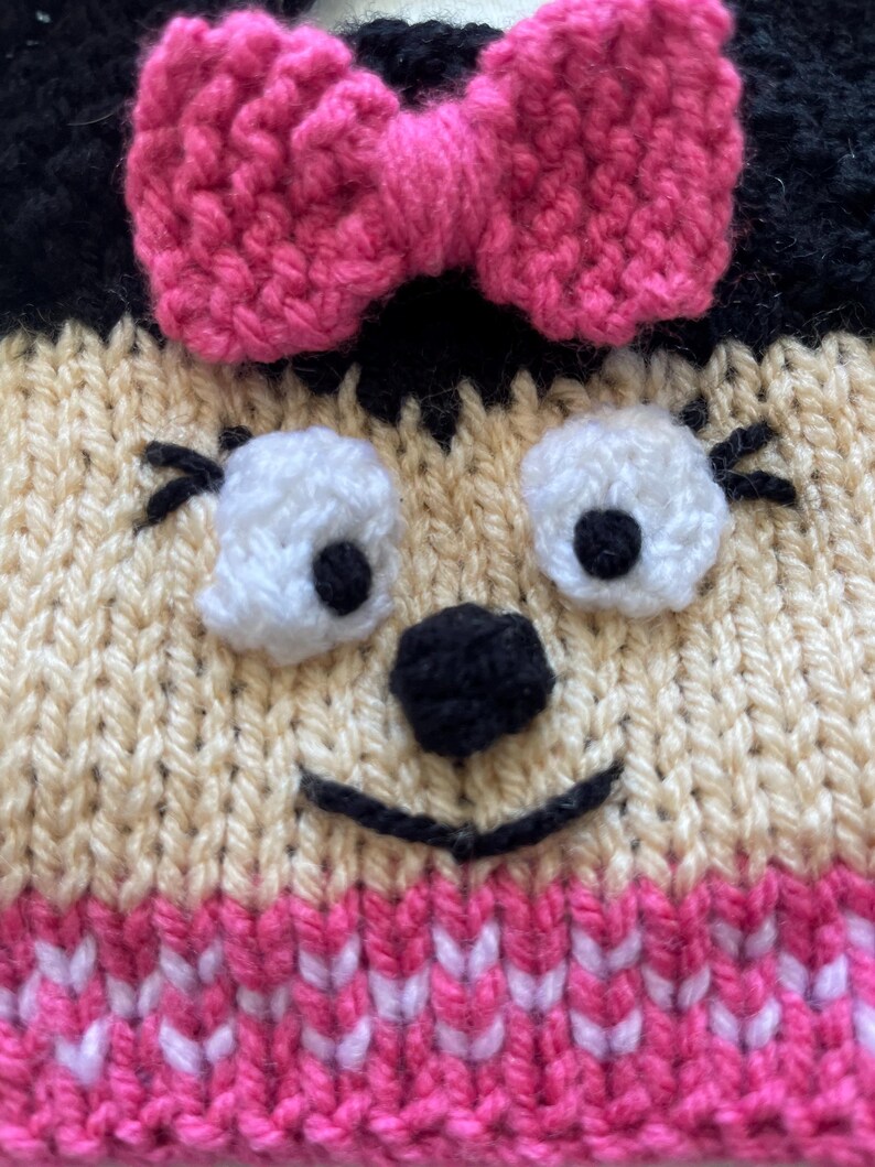 Pu&ograve; includere: Un cappello di lana con un fiocco rosa, orecchie nere e un viso da cartone animato con occhi bianchi, un naso nero e una bocca sorridente.