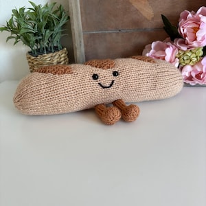 May include: A knitted plush baguette toy with a smiling face and small feet. The bread is tan with darker brown patches, and the eyes and mouth are black. The toy is on a white surface, with a small plant and flowers in the background.