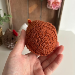 May include: A hand-knitted chicken in shades of orange and brown, with a small yellow and red comb. The chicken is held in a hand, with other knitted items and flowers in the background.