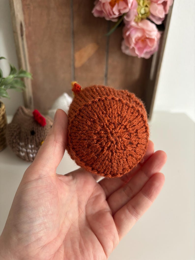 May include: A hand-knitted chicken in shades of orange and brown, with a small yellow and red comb. The chicken is held in a hand, with other knitted items and flowers in the background.
