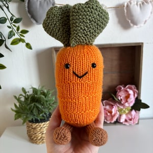 Peut inclure: Une peluche en forme de carotte tricotée à la main, de couleur orange avec des feuilles vertes et des pieds marron. La carotte a un visage souriant avec des yeux noirs et une bouche cousue. La peluche est tenue dans une main.