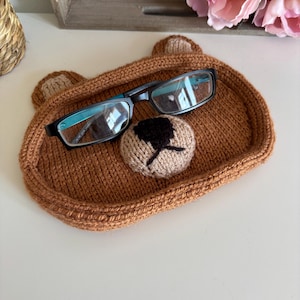 Può includere: Un vassoio lavorato a maglia di colore marrone a forma di muso d'orso, con due piccole orecchie, un muso beige chiaro e un naso ricamato in marrone scuro. Un paio di occhiali con montatura nera e dettagli color ottanio poggia sul vassoio.