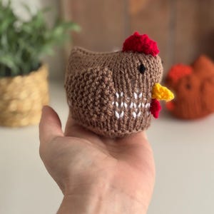 May include: A brown knitted chicken with a red comb and yellow beak is held in a hand. The chicken has white detailing on its chest and a small black eye. Another orange knitted chicken is visible in the background.