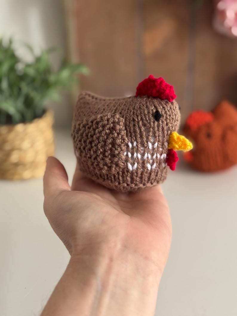May include: A brown knitted chicken with a red comb and yellow beak is held in a hand. The chicken has white detailing on its chest and a small black eye. Another orange knitted chicken is visible in the background.