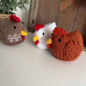 May include: Three knitted chicken figurines in shades of brown, white, and orange. Each chicken has a red comb, yellow beak, and black eyes. The chickens are on a white surface.