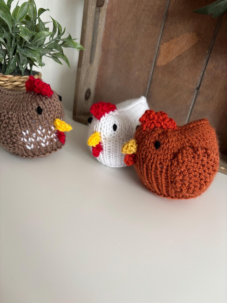 May include: Three knitted chicken figurines in shades of brown, white, and orange. Each chicken has a red comb, yellow beak, and black eyes. The chickens are on a white surface.