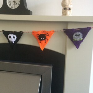 May include: A knitted Halloween banner with four triangular flags. The flags are black, orange, purple, and black. The black flags have a white skull and crossbones and a white ghost. The orange flag has a black spider. The purple flag has a white tombstone with the word "RIP" on it.