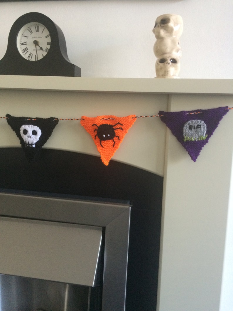 May include: A knitted Halloween banner with four triangular flags. The flags are black, orange, purple, and black. The black flags have a white skull and crossbones and a white ghost. The orange flag has a black spider. The purple flag has a white tombstone with the word "RIP" on it.