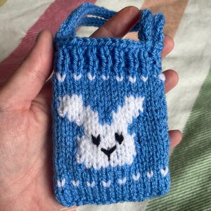 May include: A small blue knitted phone case with a white knitted dog face design. The case has a strap for carrying.