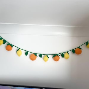 May include: A knitted garland with alternating orange and yellow fruit, resembling lemons and oranges, with green leaves. The garland is strung on a white wall.