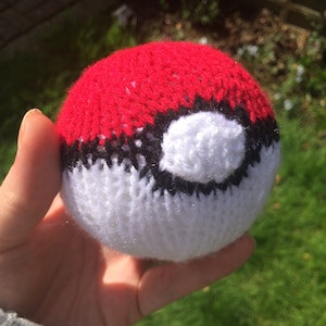 May include: A red and white knitted Poké Ball with a black centre circle. The Poké Ball is a popular item in the Pokémon franchise.