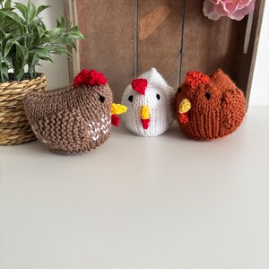 May include: Three knitted chicken figurines in brown, white, and orange. Each chicken has a red comb and yellow beak. The chickens are handmade and sit on a white surface. A small potted plant and a wooden tray with pink flowers are in the background.
