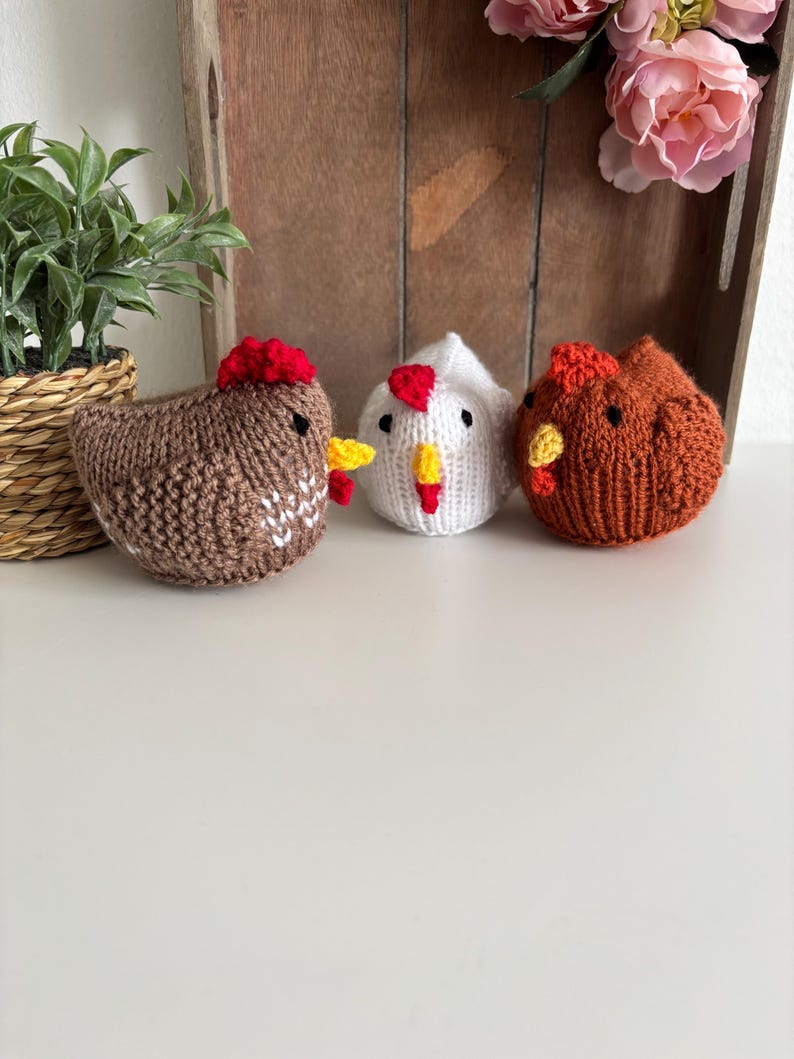 May include: Three knitted chicken figurines in brown, white, and orange. Each chicken has a red comb and yellow beak. The chickens are handmade and sit on a white surface. A small potted plant and a wooden tray with pink flowers are in the background.