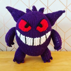 Puede incluir: Una criatura tejida a mano de color púrpura con ojos rojos y una amplia sonrisa blanca. El peluche tiene orejas puntiagudas y brazos y piernas pequeños. El juguete está sobre una superficie de madera.