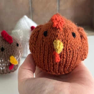 May include: Close-up of three knitted chicken figures. The front chicken is orange with a red comb and yellow beak. The other two chickens are brown and white, also with red combs and yellow beaks. The chickens are handmade and appear soft.