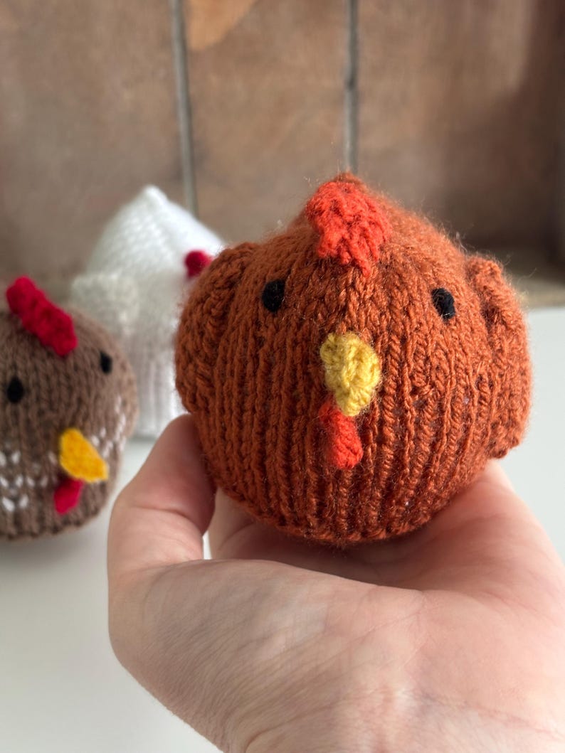 May include: Close-up of three knitted chicken figures. The front chicken is orange with a red comb and yellow beak. The other two chickens are brown and white, also with red combs and yellow beaks. The chickens are handmade and appear soft.