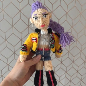 May include: A handmade knitted doll with purple hair styled in a high bun and a long braid. The doll wears a yellow jacket with colorful patches, denim shorts, and black boots with red stripes. The doll has a silver necklace and earrings.