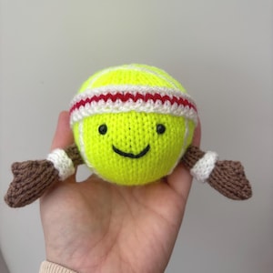 Könnte beinhalten: Ein gestricktes Tennisball-Spielzeug mit einem lächelnden Gesicht, schwarzen Augen und einem schwarzen, aufgenähten Mund. Der Tennisball trägt ein weiß-rotes Stirnband und braune Arme mit weißen Manschetten. Das Spielzeug ist gelb und weiß.