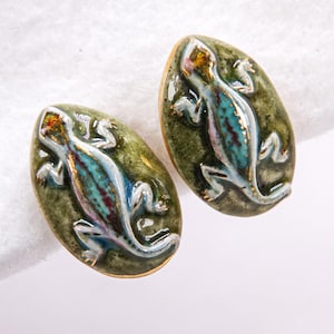 May include: A pair of ceramic earrings. Each earring is oval-shaped with a green background and a detailed lizard design in shades of blue, red, and gold. The edges of the earrings are trimmed with gold.