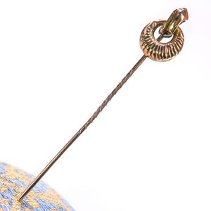 May include: A gold-coloured hat pin featuring a decorative ring-shaped head and a twisted shaft. The pin is angled, with the point inserted into a blue and yellow patterned fabric.