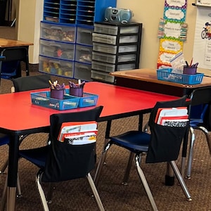 May include: A classroom scene featuring a red table with blue chairs. Black fabric pouches hang on the chair backs, holding notebooks and papers. Blue storage containers and a small radio sit in the background, suggesting an educational environment.