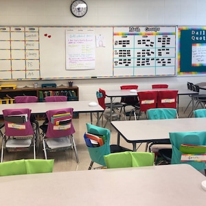 May include: A classroom setting with desks, chairs, and whiteboards. Chairs have colorful pockets labeled with numbers. Whiteboards display "Math Groups" and "All about me!" notes. A clock hangs on the wall.