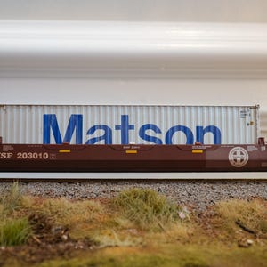 Op de afbeelding: Een modeltreinwagon met een container. De container is wit met het blauwe woord "Matson" erop gedrukt. De treinwagon is bruin met de tekst "BNSF 203010" erop gedrukt. De treinwagon staat op een spoor.