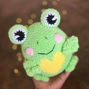 Puede incluir: Peluche de rana de ganchillo verde y redondo, hecho a mano, con grandes ojos con borde blanco, mejillas rosas y vientre amarillo. La rana tiene brazos y piernas pequeños y una cara sonriente. Un juguete suave y lindo.