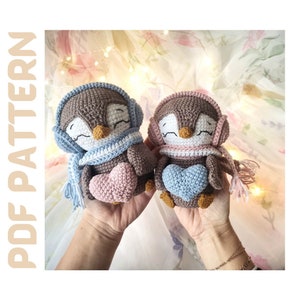 Op de afbeelding: Twee gehaakte amigurumi-pinguinspeelgoedjes met bruine lichamen, witte en blauwe sjaals en roze en blauwe harten. De pingu&iuml;ns houden de harten in hun flippers.