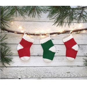 May include: Three miniature crocheted Christmas stockings hanging on a branch with white lights. The stockings are red, green, and white.
