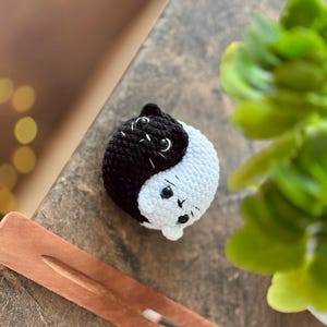 Pode incluir: Um brinquedo de gato de croch&ecirc; preto e branco em forma de s&iacute;mbolo yin yang. O gato tem olhos pretos e um nariz branco.