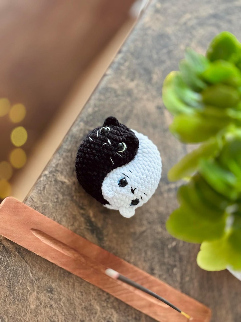 Pode incluir: Um brinquedo de gato de croch&ecirc; preto e branco em forma de s&iacute;mbolo yin yang. O gato tem olhos pretos e um nariz branco.